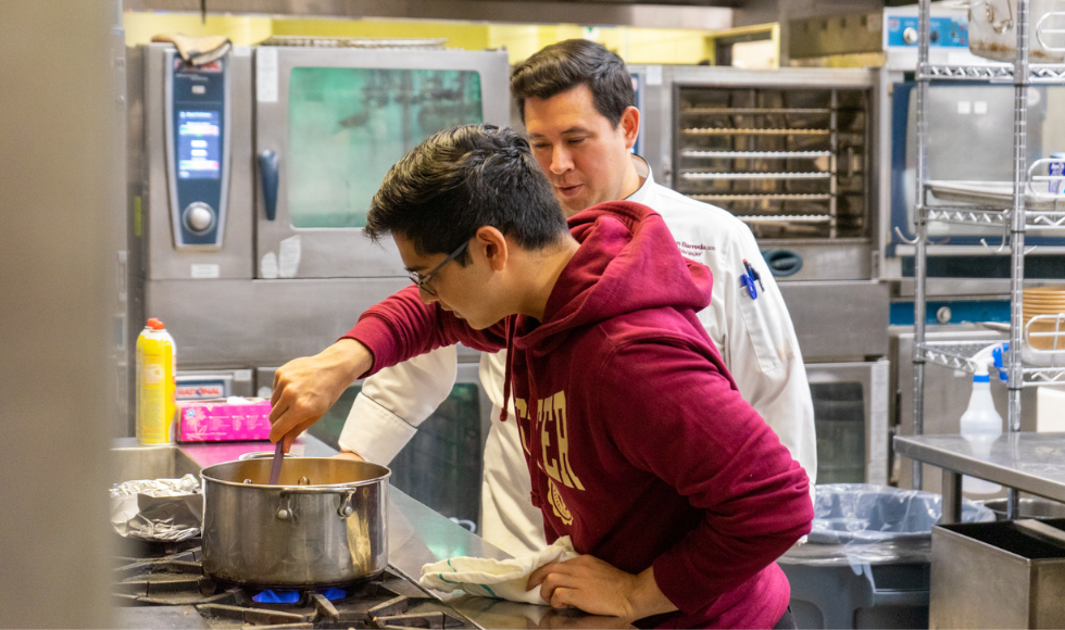 Student stirring a pot on a gas range with chef standing in observation