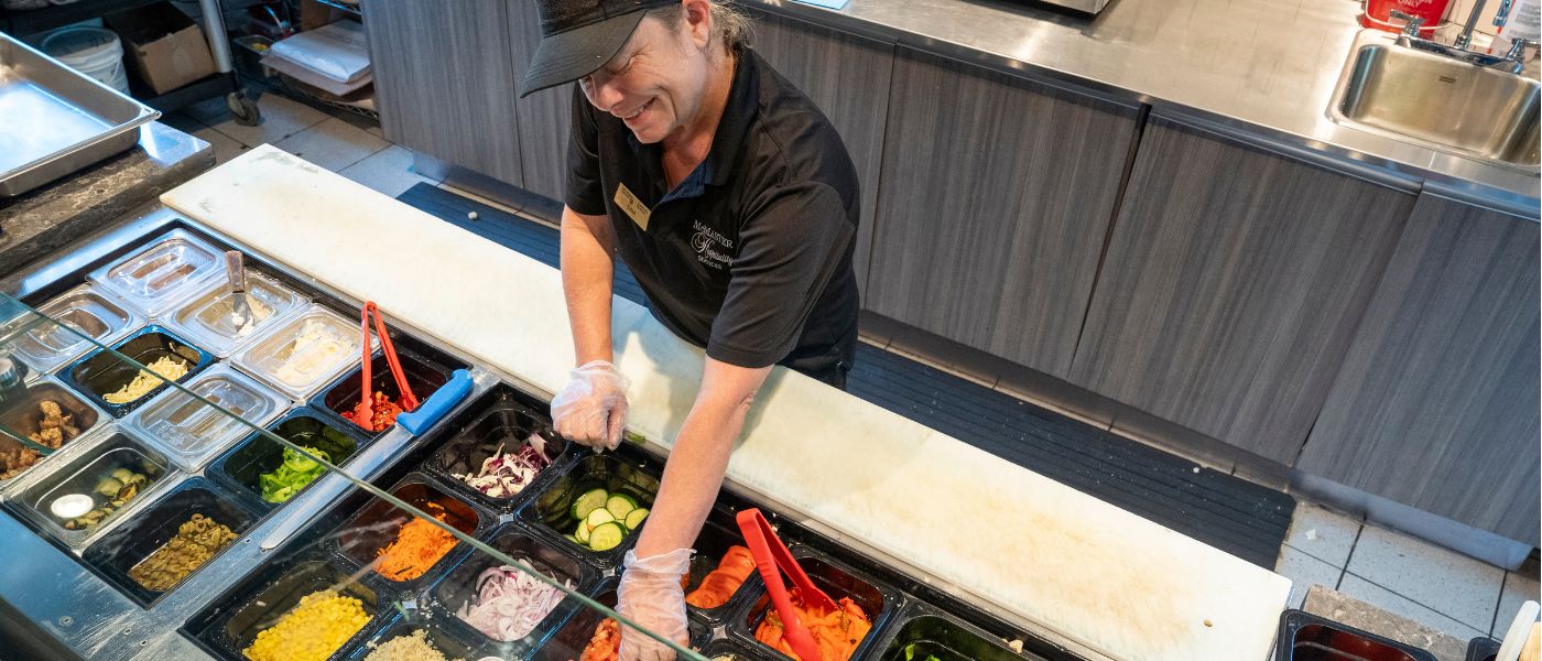 Hospitality employee reaching for veggies at a sandwich prep station with several trays of assorted veggies